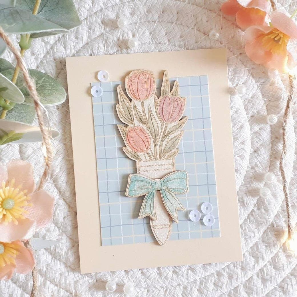 Carte crème : un bouquet de tulipes roses et corail noué d'un ruban bleu, découpé et aquarellé, sur un fond à petits carreaux.